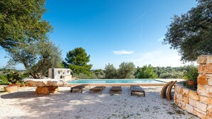 A pool area with a stone bench, lounge chairs, and a view of trees and hills. - Holiday Home Trullo with Shared Pool, Shared Garden & Terrace (Ostuni)