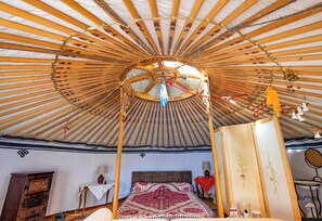 L’intérieur d’une yourte traditionnelle présente une coupole centrale, un lit avec une literie à motifs, un meuble en bois et une petite table avec une lampe.