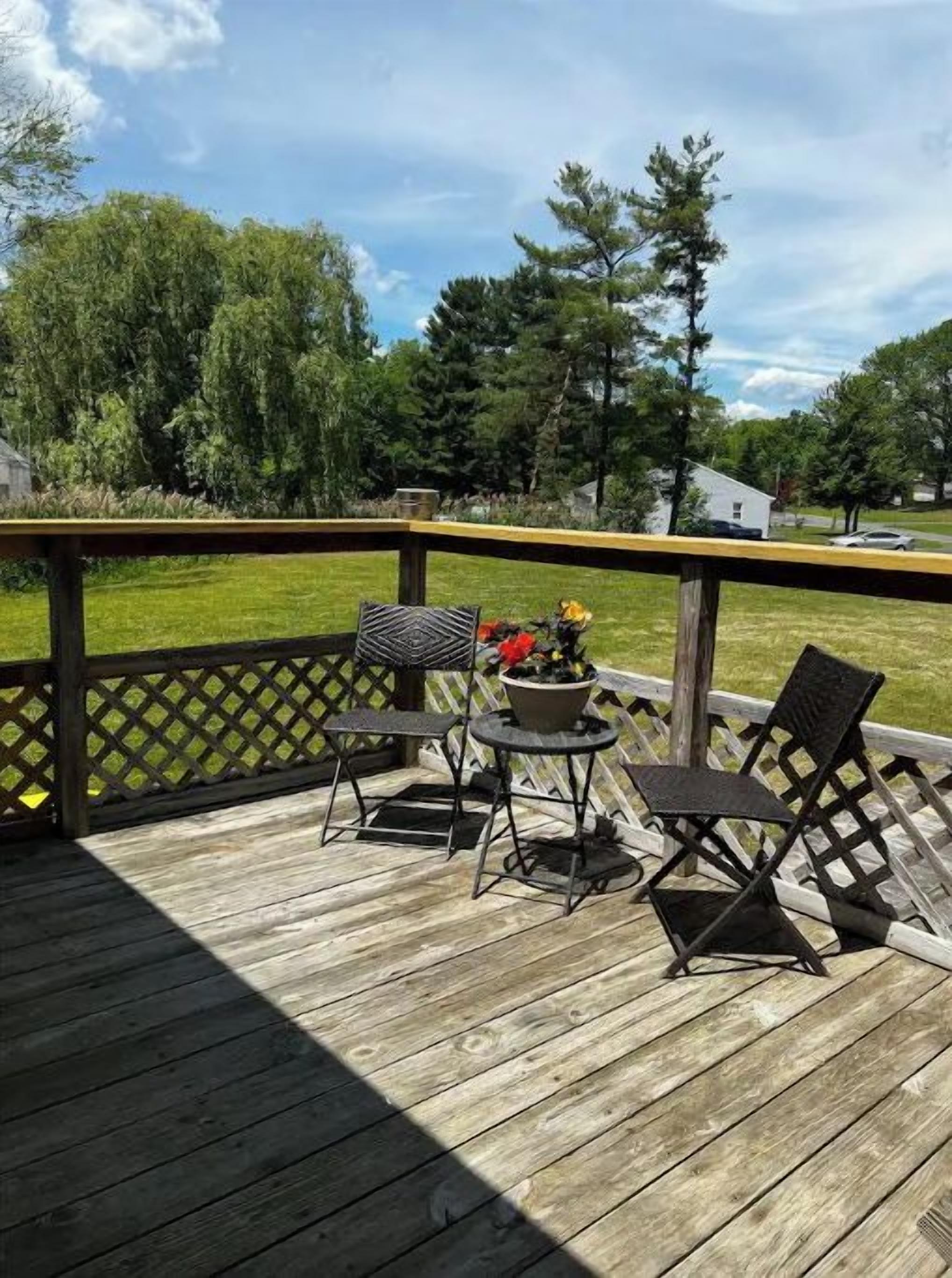 Eine Holzterrasse mit Gittergeländer, zwei Metallstühlen, einem kleinen runden Tisch und einem Blumentopf.