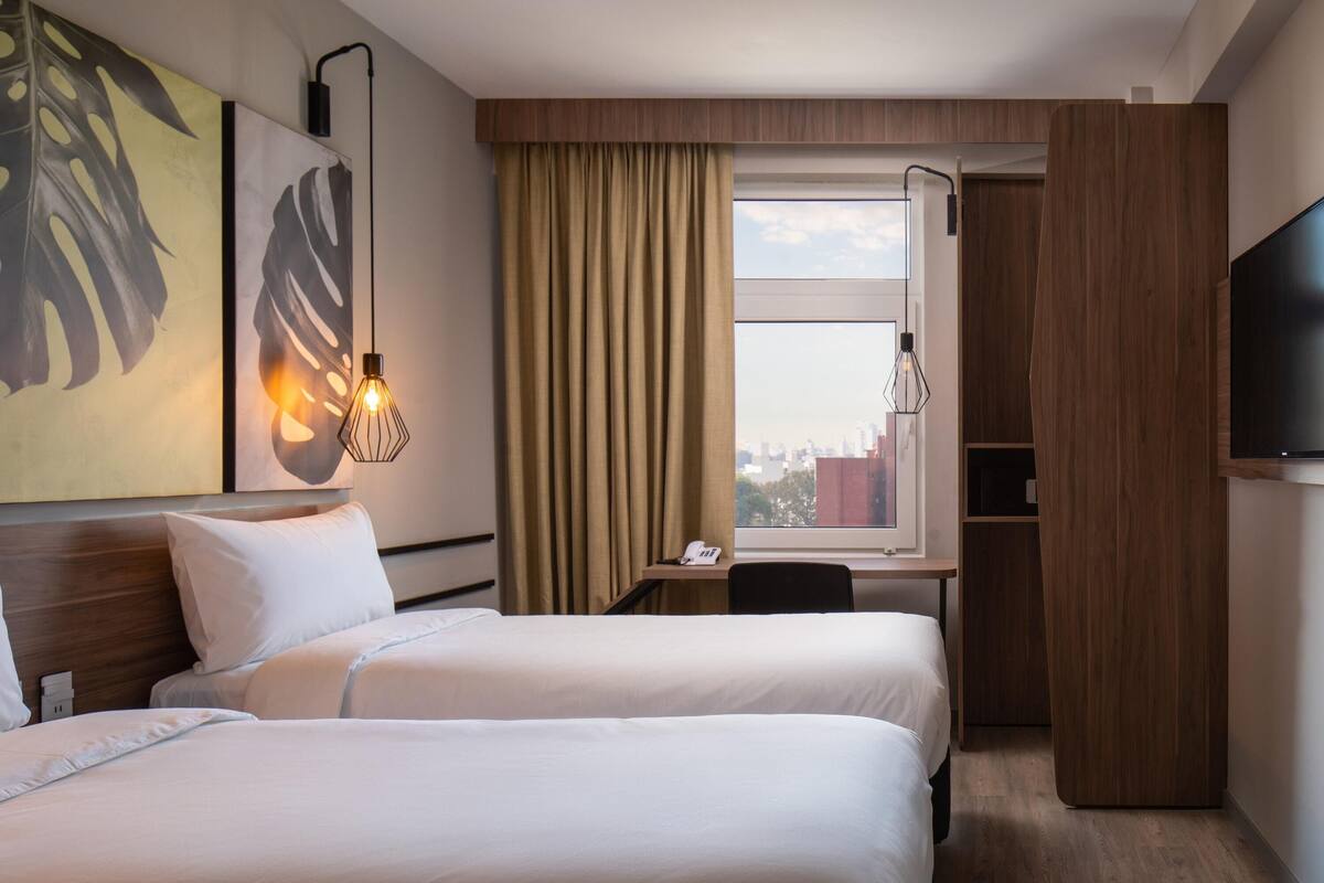 Een hotelkamer met twee bedden, een bureau, een stoel, een televisie, een raam met gordijnen en kunst aan de muur.