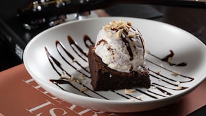 Un dessert servi dans une assiette, nappé de sauce au chocolat et agrémenté de noix, avec une guitare en arrière-plan.