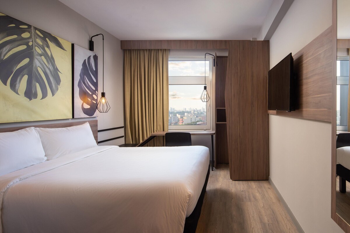 Une chambre d’hôtel avec un grand lit, un bureau, une chaise, une télévision et une fenêtre avec des rideaux.