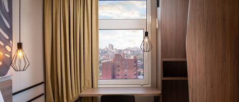 A hotel room with a bed, a desk with a chair, a view of the city through a window, and two pendant lights.