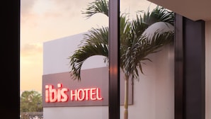 L’intérieur d’un hôtel au design moderne, avec une grande fenêtre donnant sur des palmiers et une enseigne indiquant « ibis HOTEL ». On y trouve trois chaises vertes et une petite table ronde.