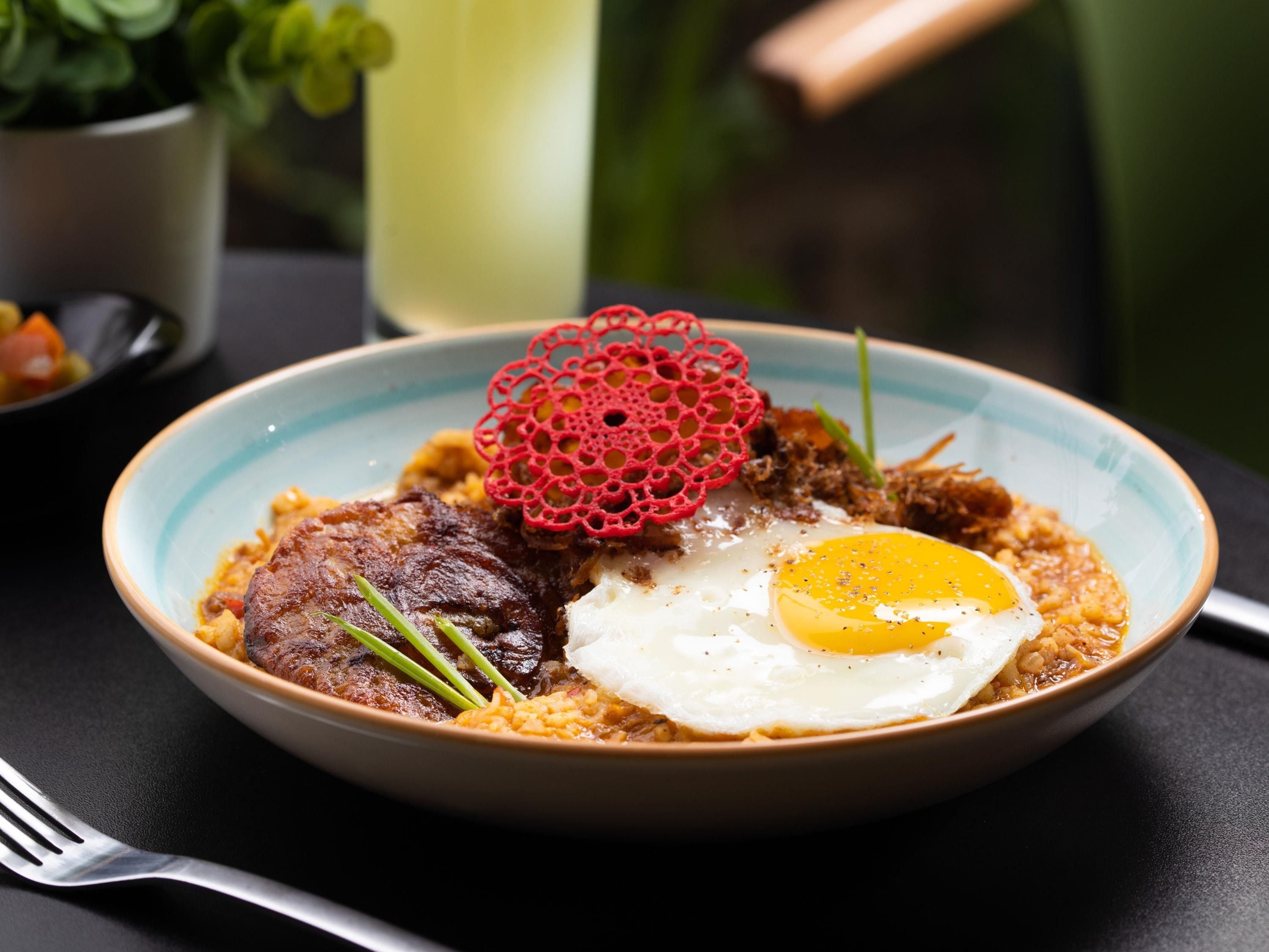 A plate with a fried egg, a piece of meat, and a decorative red item on a table.