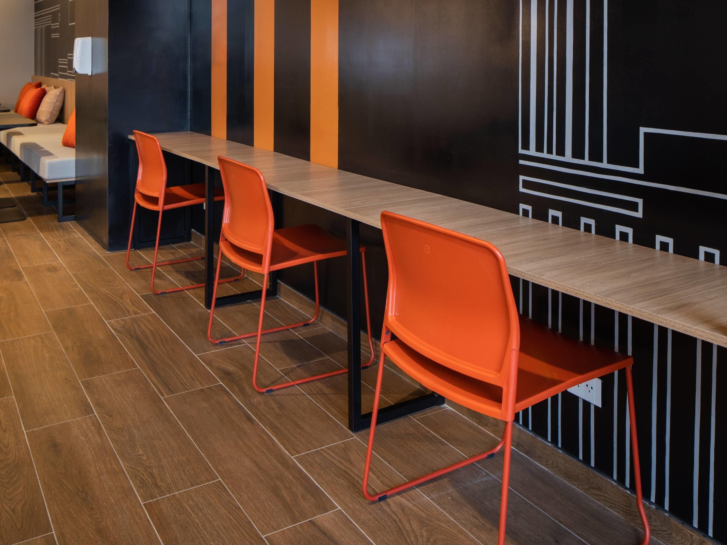 A modern hotel lobby with a long wooden reception desk, orange chairs, and a black and orange striped wall.