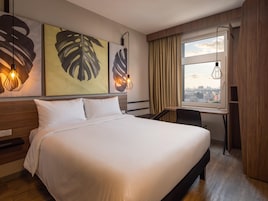 Una habitación de hotel con una cama grande, un escritorio, una silla, un ventanal con cortinas y una obra de arte en la pared.