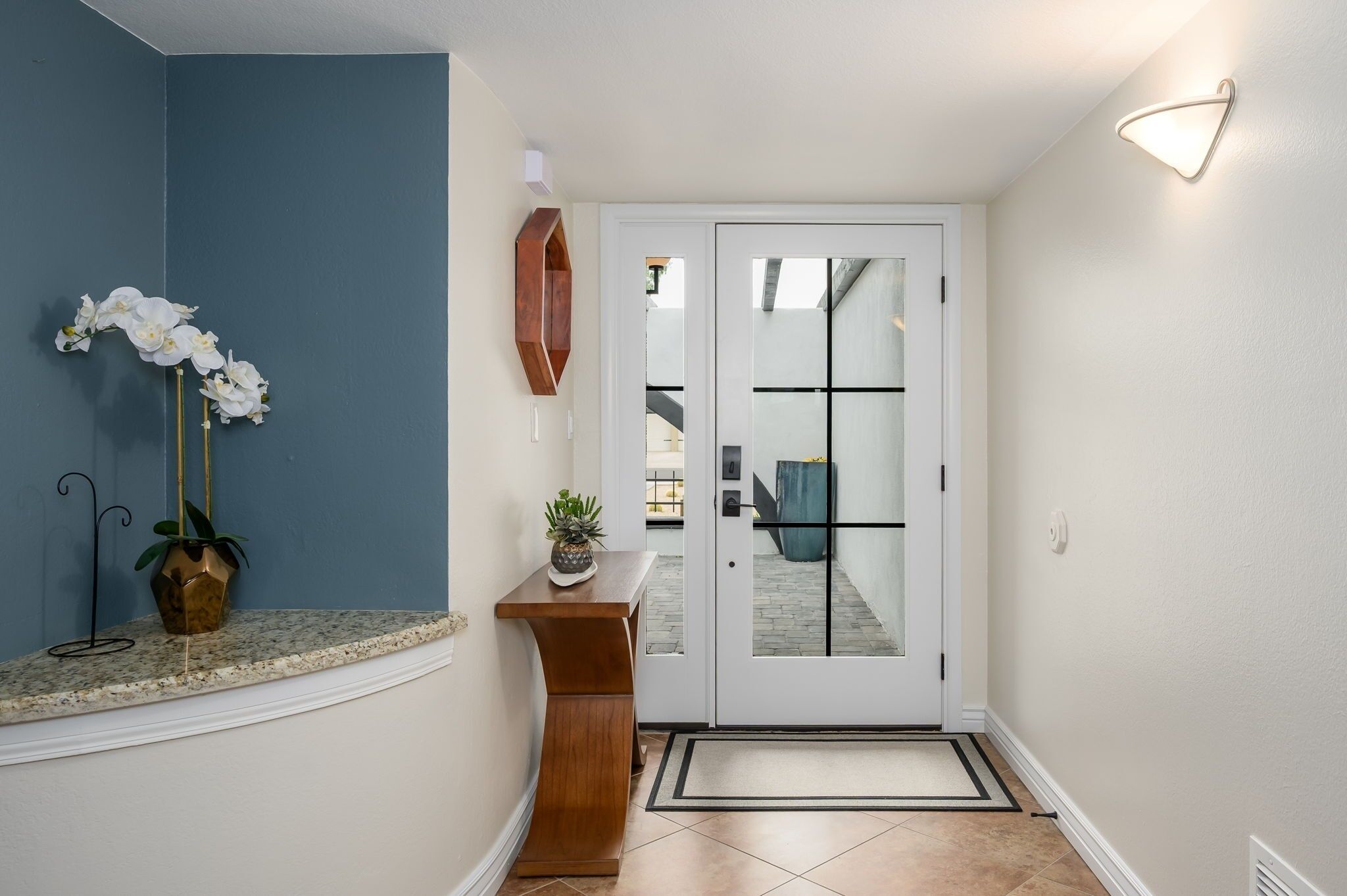 A hallway with a blue wall, a wooden bench with a potted plant, a white door with glass panels, and a light fixture on the wall.
