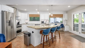 Uma cozinha moderna com armários brancos, eletrodomésticos de aço inoxidável, bancada de granito e uma ilha central com banquetas.