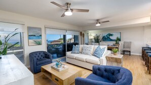 A spacious living room with a white sofa, blue armchairs, and a wooden coffee table. There are ceiling fans, a view of the ocean through large windows, and a dining area with chairs. - Kaanapali Plantation 3 bedroom 2.5 baths OceanView 3 Condo by RedAwning (Lahaina)
