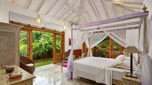 Tilava makuuhuone, jossa on suuri sänky, oleskelualue pöydällä ja tuoleilla sekä avoimesta ovesta avautuva näkymä vehreään maisemaan.