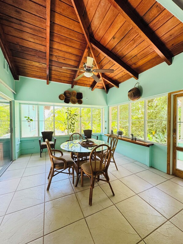 A dining area with a round table and chairs, a ceiling fan, and large windows with a view of greenery outside. - Caribbean Villa with private pool & ocean view in the heart of Montserrat . (Woodlands)