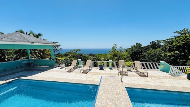 A clear blue pool with a tiled deck, lounge chairs, and a view of the ocean.