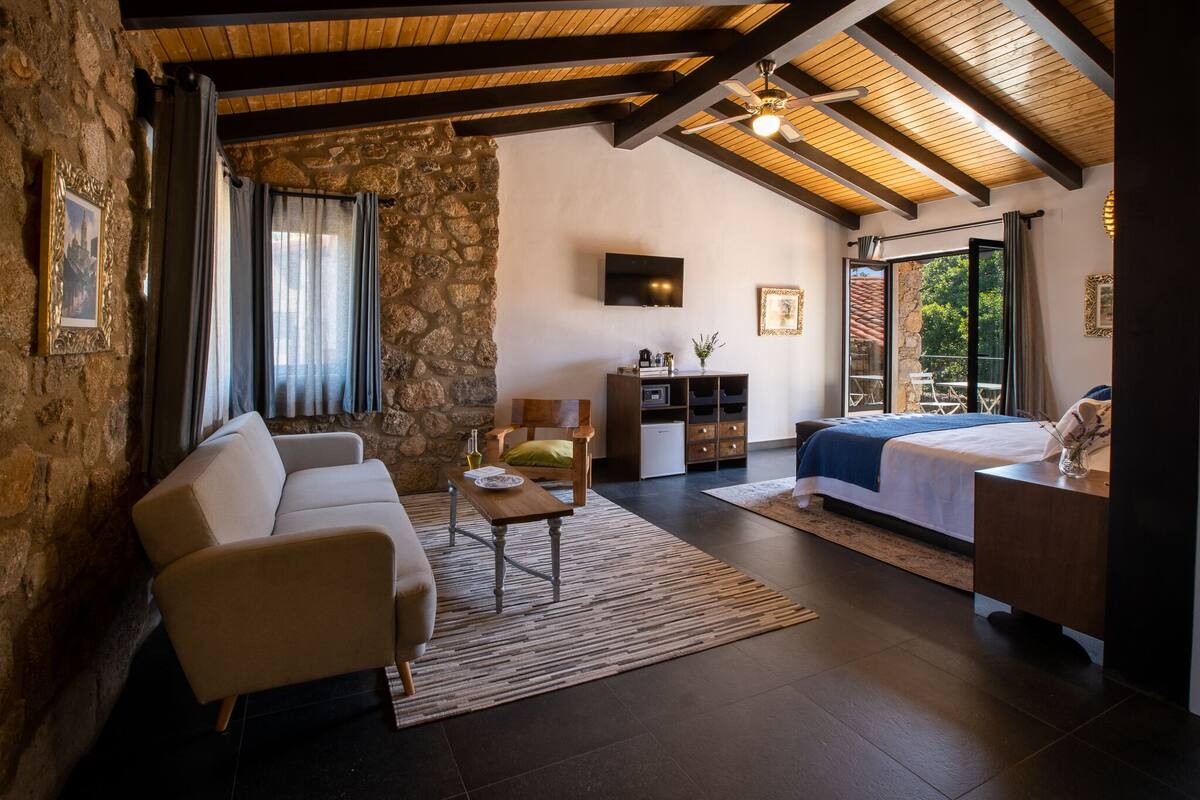 Ein gemütlicher Raum mit Steinwand, Holzbalkendecke, einem Bett, einer Couch, einem Couchtisch und einem Fernseher.