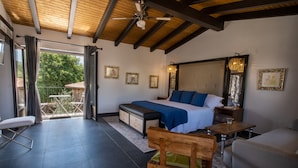 Een slaapkamer met een groot bed, een houten bankje, een bureau en een stoel. Er is een balkon met een tafel en stoelen, en uitzicht op bomen.