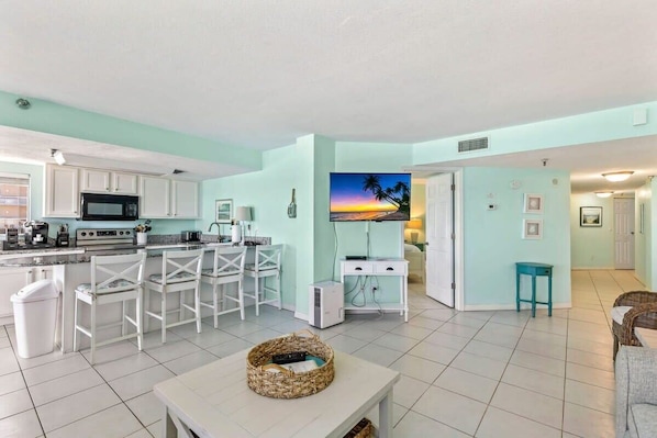 A living room with a TV, kitchen area with white cabinets, and a dining area with white chairs.