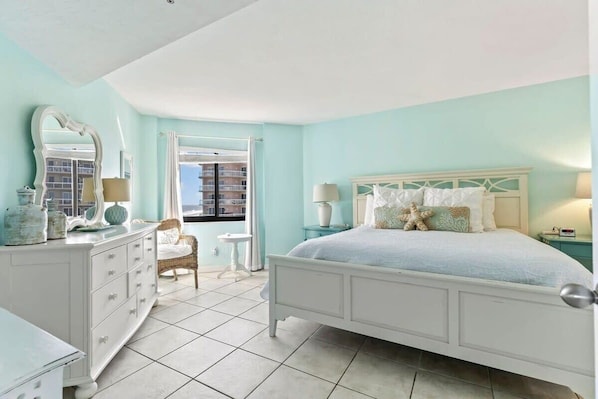Un dormitorio con cama, cómoda y una silla. Se ve una ventana con vista a edificios.