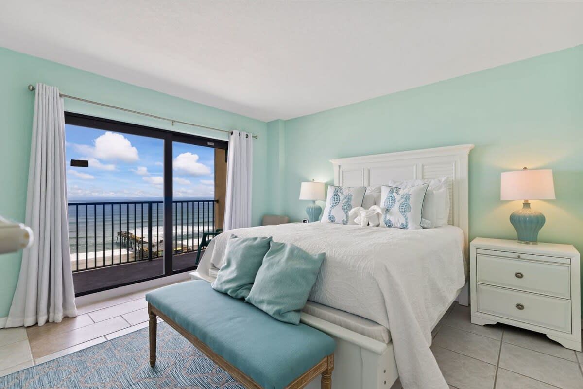 A bedroom with a large bed, a bench, a nightstand with a lamp, and a view of the ocean through a sliding door.