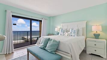 A bedroom with a large bed, a bench, a nightstand with a lamp, and a view of the ocean through a sliding door.