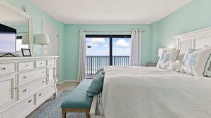 Une chambre avec un grand lit, une commode, un banc et une vue sur l’océan depuis un balcon.