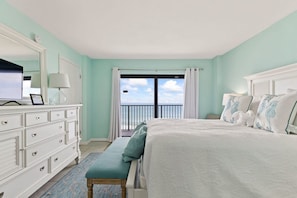 A bedroom with a large bed, a dresser, a bench, and a view of the ocean through a balcony.