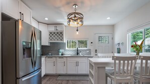 A modern kitchen with white cabinetry, a stainless steel refrigerator, a dining area with a white table and chairs, and a view of greenery outside. - Kailua Beach/Lanikai LUX apartment w/Ocean Views from Rooftop Sun Deck (Kailua)