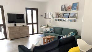 A living room with a dark blue sofa, a wooden coffee table, and a yellow chair. There is a TV on a wooden cabinet, and a wall decorated with framed pictures. - Villa del Brigante, 200m to the beach, 80m² (Quartu Sant'Elena)