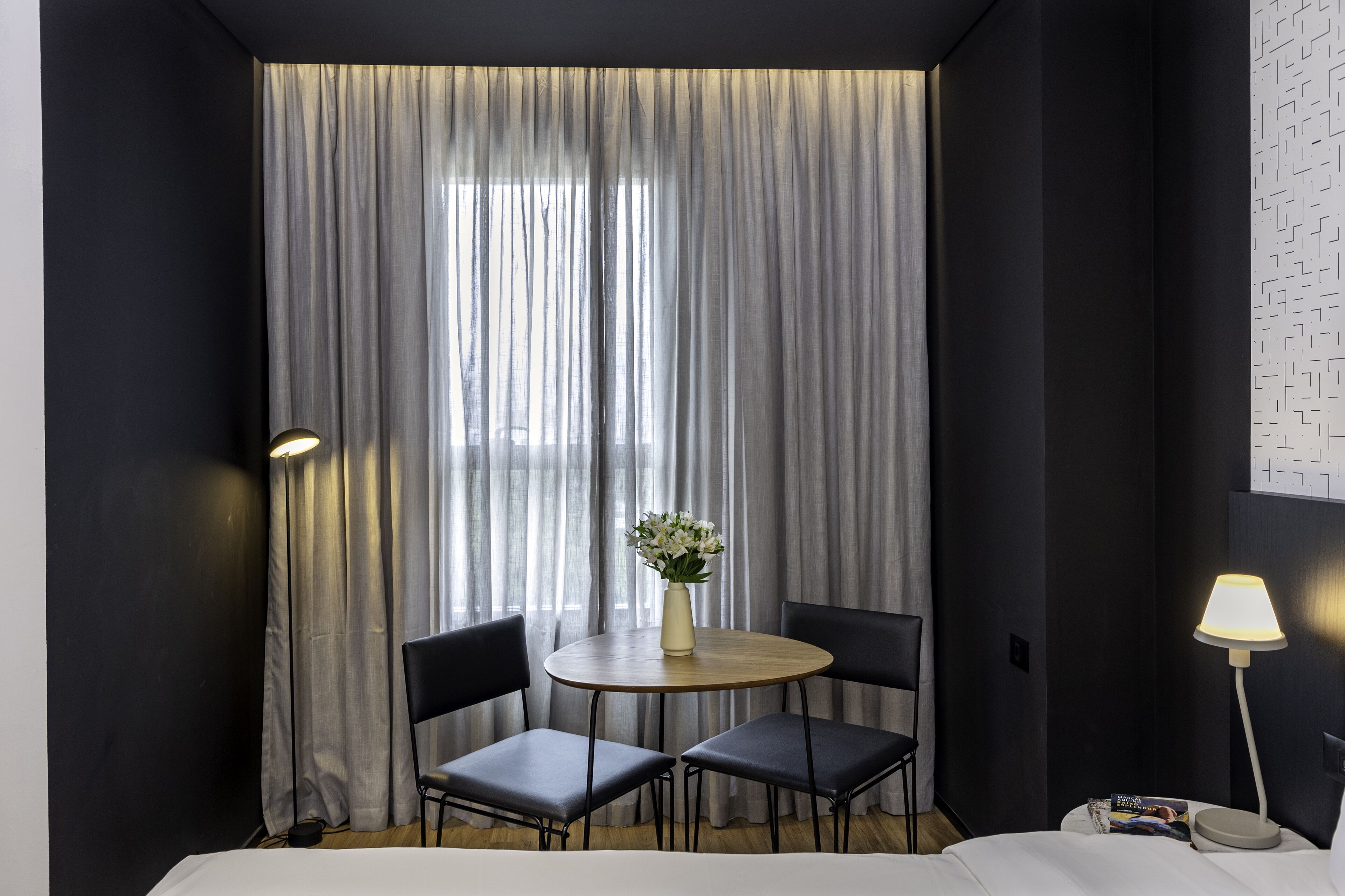 A hotel room with a round table, two chairs, a vase with flowers, and a lamp on a side table.