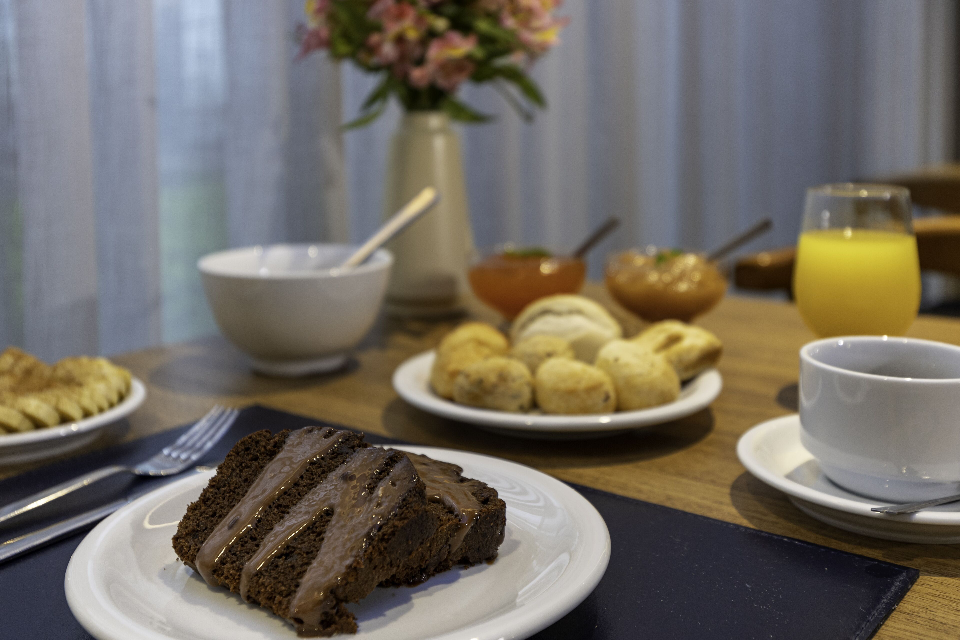 A breakfast table with a slice of chocolate cake, a bowl of fruit, a cup of coffee, and a glass of orange juice.