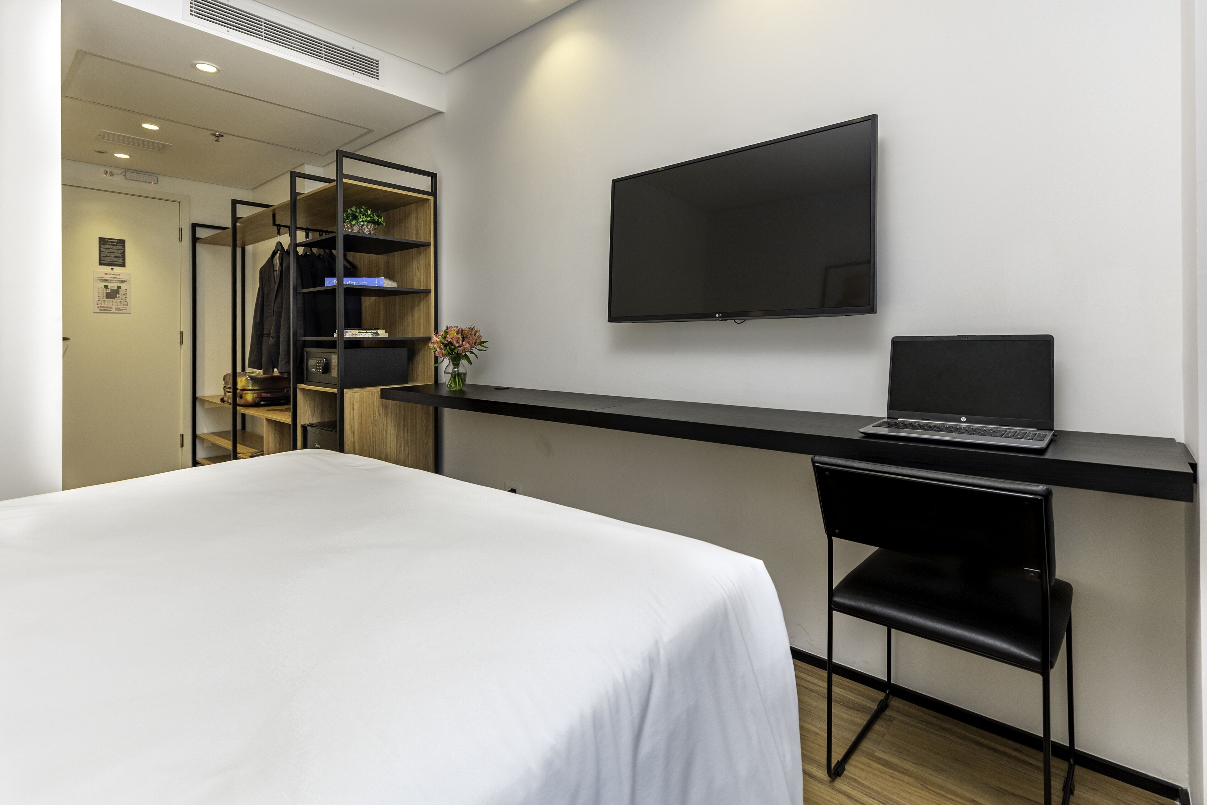 A hotel room with a bed, a desk with a laptop, a chair, a TV, and a wardrobe.