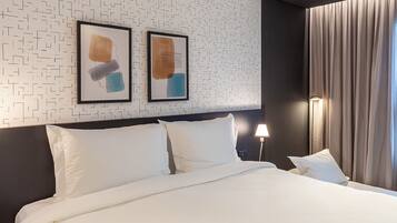 A hotel room with a neatly made bed, two framed abstract artworks on the wall, a dark headboard, and a large window with curtains.