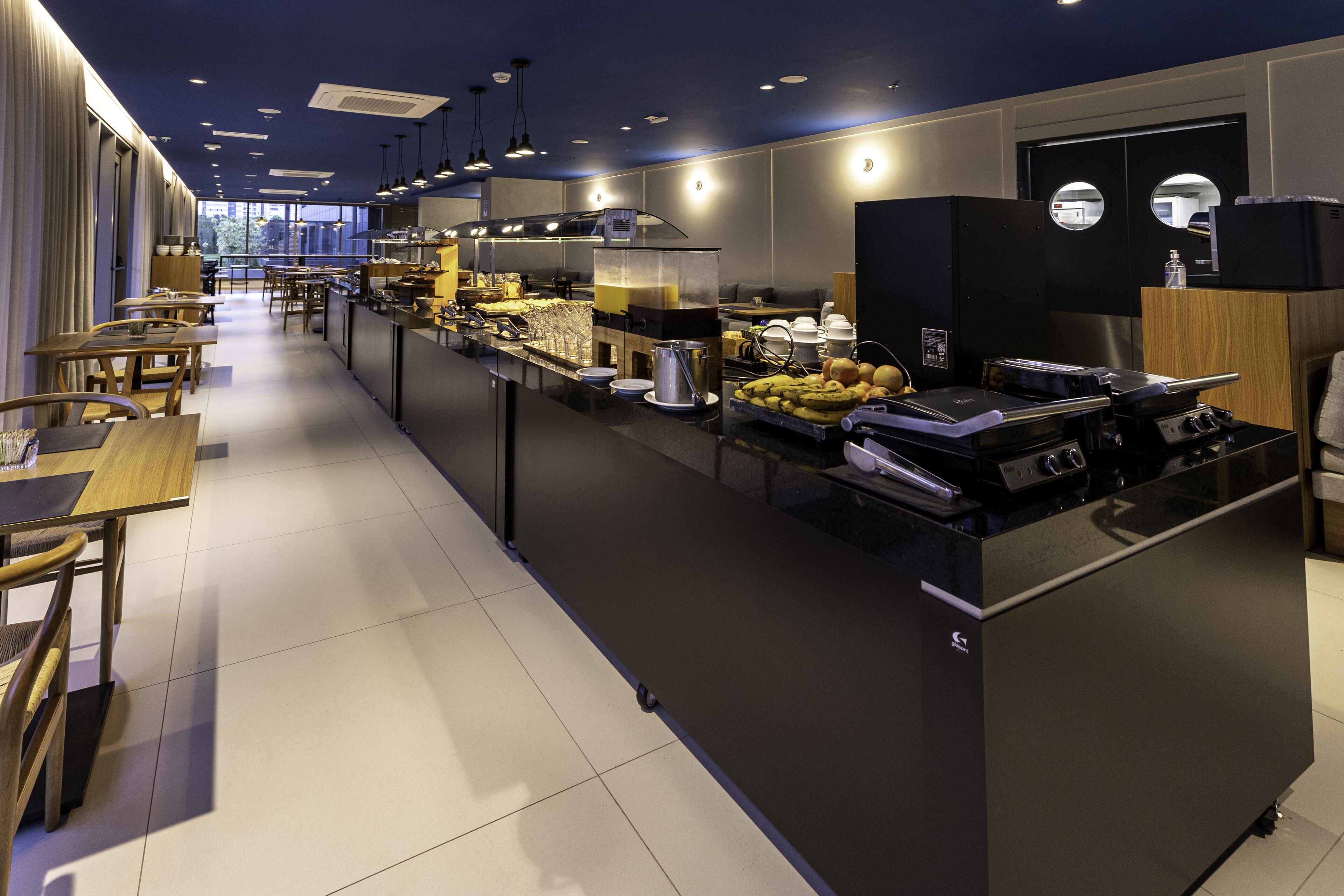 A modern hotel kitchen with a buffet, dining tables, and a view of the city.