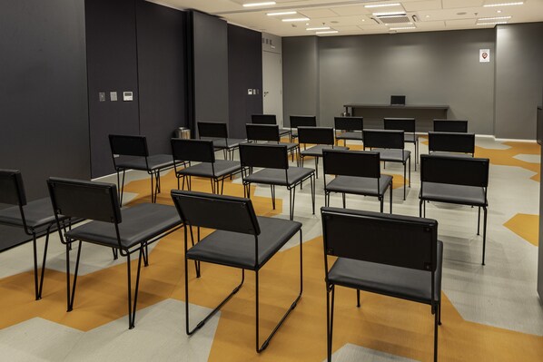 Un salón de conferencias con filas de sillas negras y mesas, un atril y un letrero de 'No Fumar' en la pared.