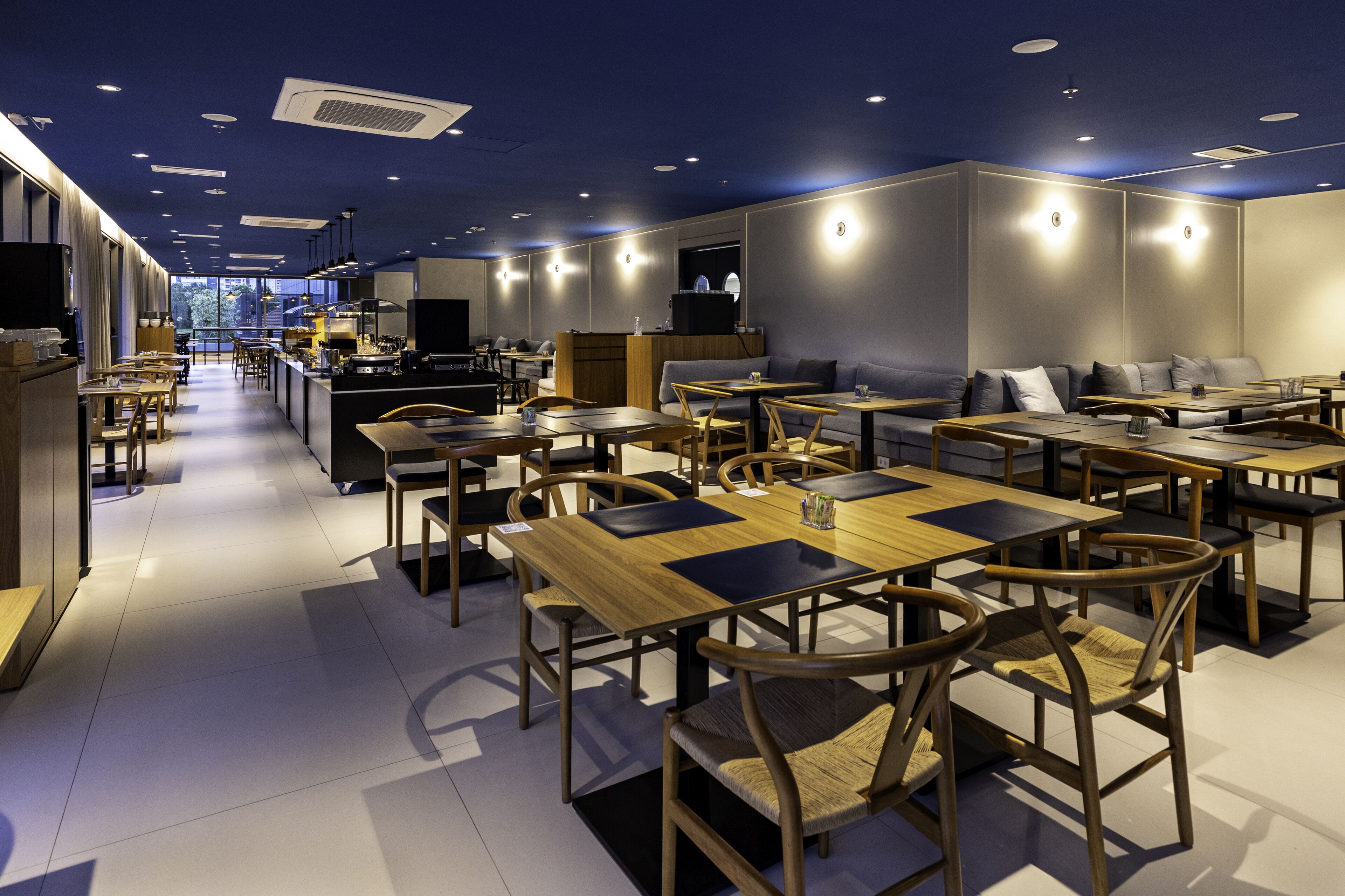 A modern restaurant with wooden tables and chairs, a bar area, and a view of the city through large windows.