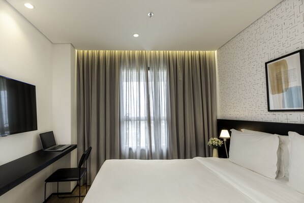 Una habitación de hotel moderna con una cama, un escritorio con una computadora portátil, una silla, una mesita de noche con una lámpara y un ventanal con cortinas.
