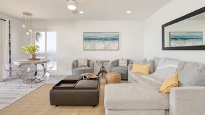 A modern living room with a grey sofa, a brown ottoman, a round coffee table, and a small side table. There is a large window with a view of the ocean, and a ceiling fan is visible. - Oceanfront in La Jolla, Steps to Beach, Shops, Dining | Dawn Patrol by AvantStay (La Jolla)