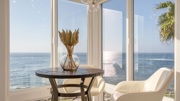 A room with a large window overlooking the ocean, a round table with a vase, and a white chair. - Oceanfront in La Jolla, Steps to Beach, Shops, Dining | Dawn Patrol by AvantStay (La Jolla)