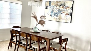 Un comedor con una mesa de madera, sillas, una araña, un jarrón con plantas secas y un cuadro en la pared.