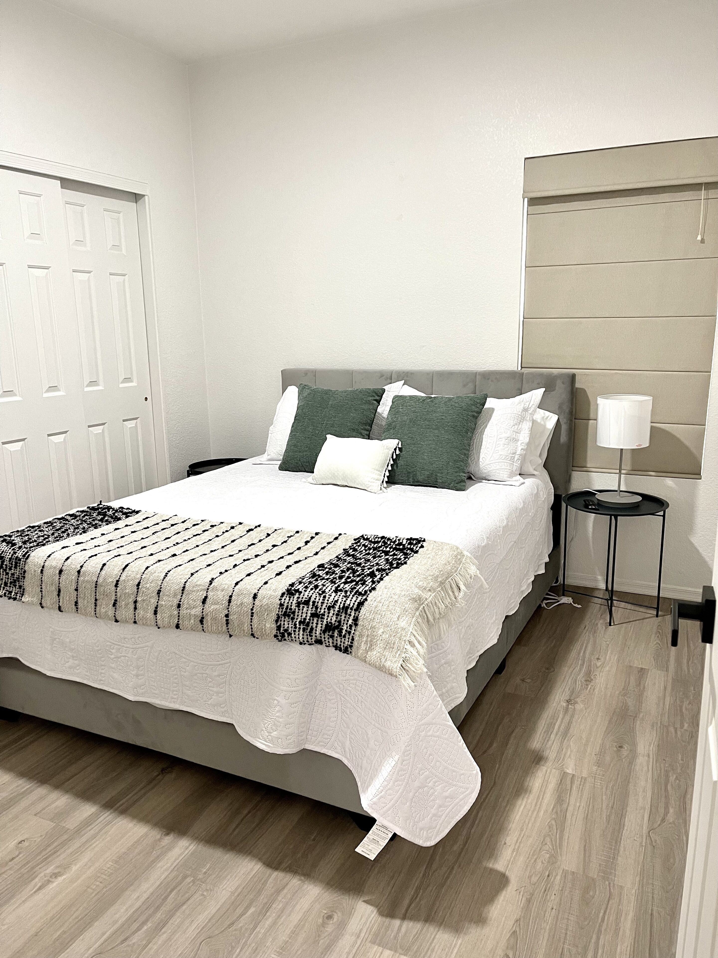 A neatly made bed with a white and black patterned bedspread and pillows, a bedside table with a lamp, and a wooden floor.