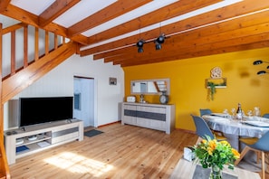 A spacious living area with a wooden staircase, a flat-screen TV, a dining table set for six, and a yellow accent wall.