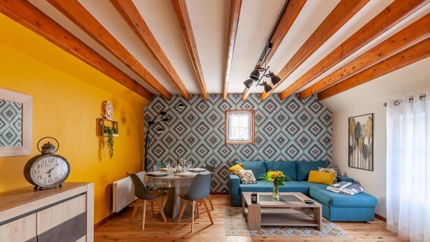 Un salon chaleureux avec des poutres en bois, un coin repas avec des chaises et une table, et un canapé bleu agrémenté de coussins.