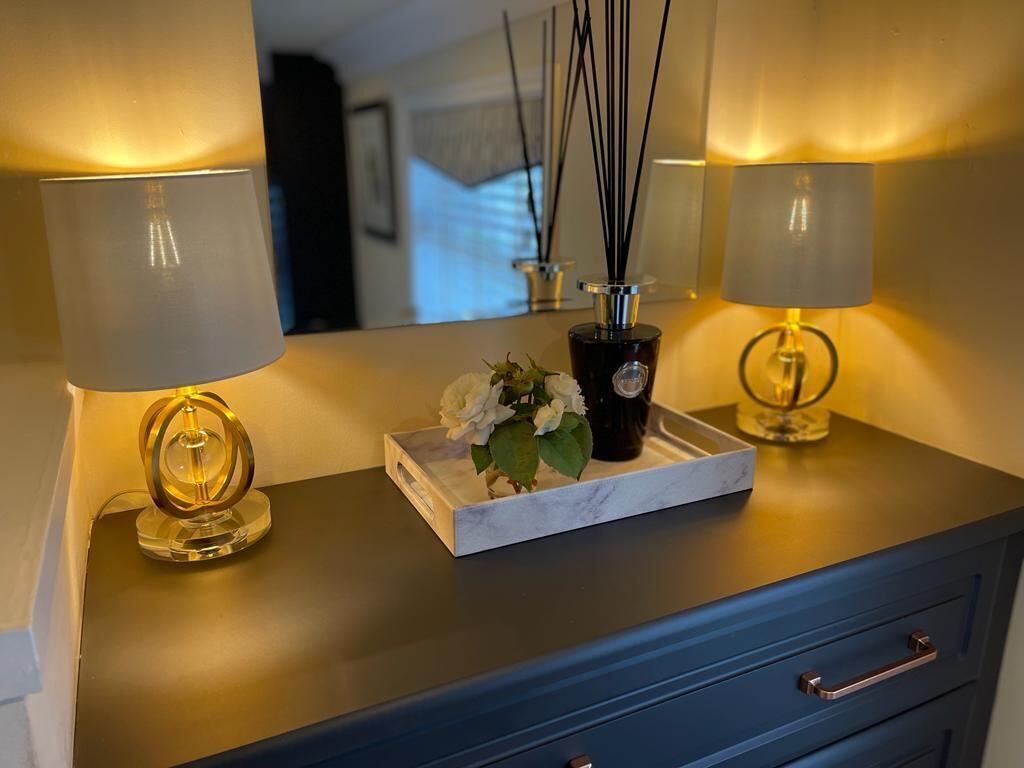 A dark blue dresser with two lamps on either side, a marble tray with a black vase and white flowers, and a mirror reflecting the room.