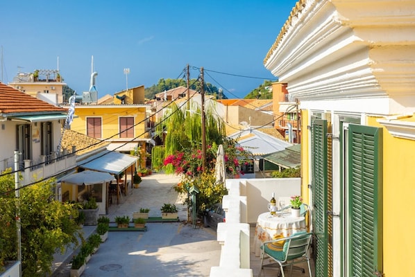 Una calle estrecha flanqueada por edificios, una pequeña mesa con una silla, y un balcón con contraventanas verdes.