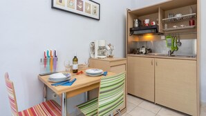 Ett litet, praktiskt kök med ett träbord, stolar och en randig handduk. En kaffemaskin och en vattenkokare står på bänkskivan.