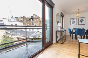 A modern apartment with a large balcony, wooden flooring, a dining area with blue chairs, and a view of the cityscape.