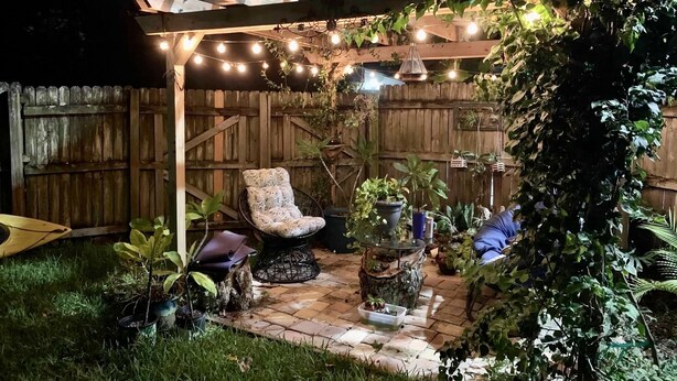 Ein Patio mit Pergola, Lichterketten, einem gepolsterten Stuhl und Kübelpflanzen.