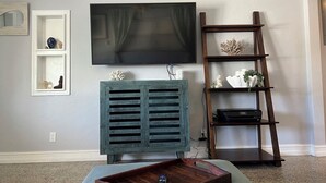 Ein Wohnzimmer mit Flachbildfernseher an der Wand, einem Holz-TV-Möbel mit Dekoration, einem Holzleiterregal mit Dekoelementen und einer Holzablage auf dem Boden.
