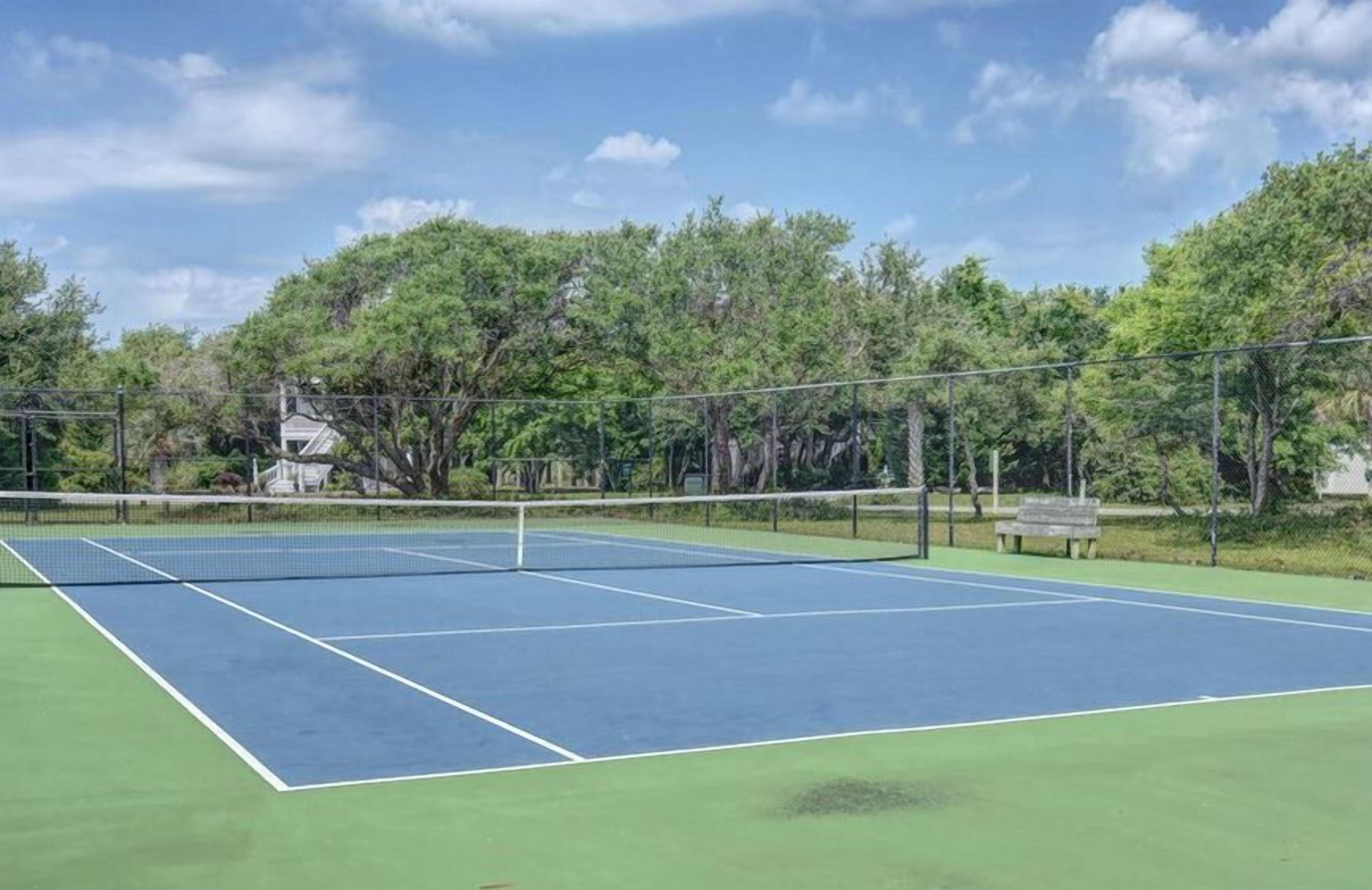 Cancha de tenis con red, rodeada de árboles y una banca.