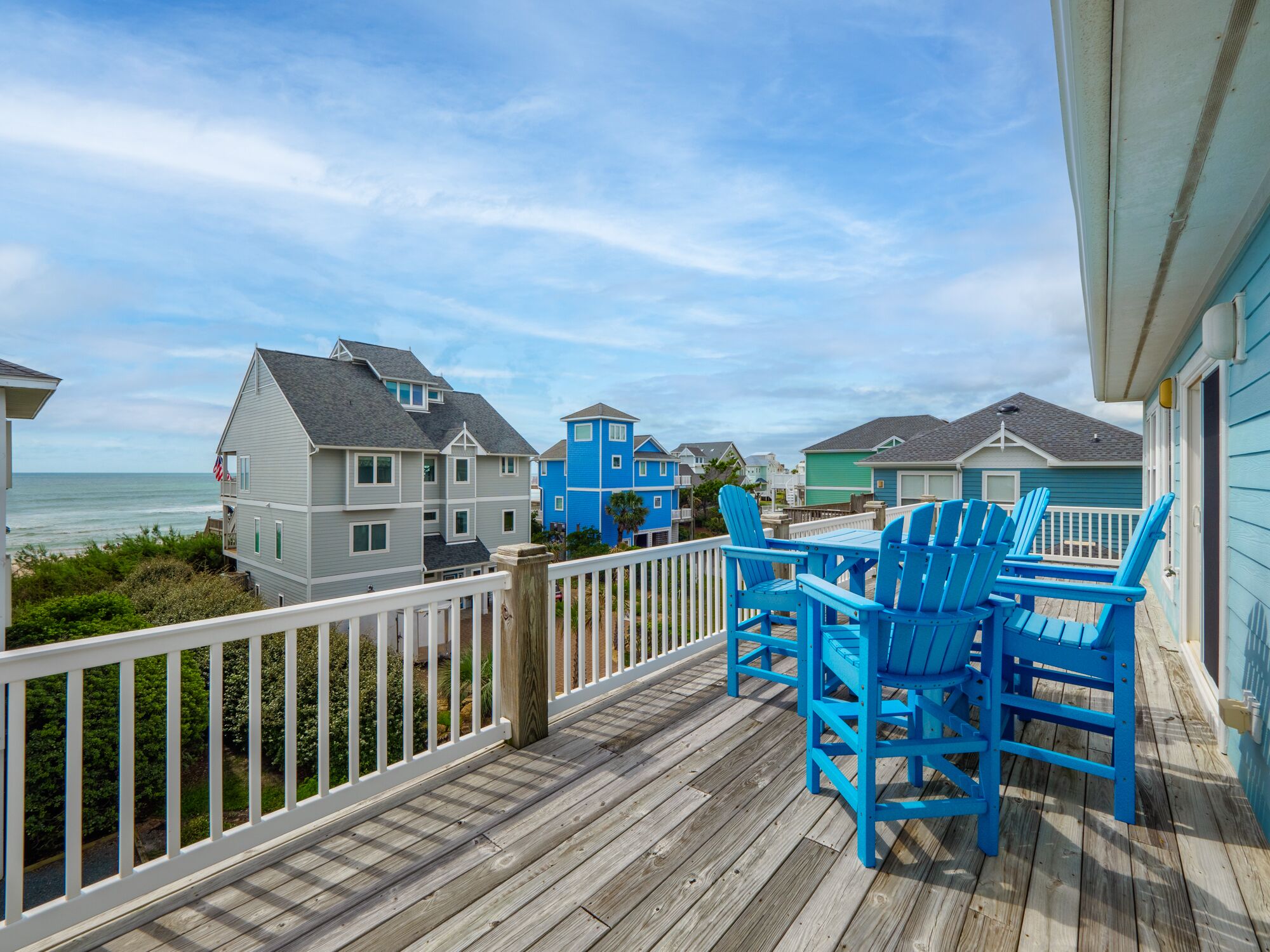 A wooden deck with blue Adirondack chairs, overlooking a beach with houses in the background.