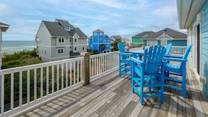 A wooden deck with blue Adirondack chairs, overlooking a beach with houses in the background. - Summer Wynd | Ocean Views + Community Amenities! (North Topsail Beach)